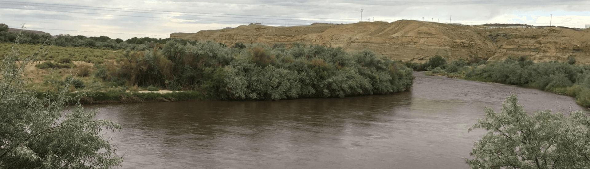 A winding river flows alongside lush greenery and rolling hills under a cloudy sky.