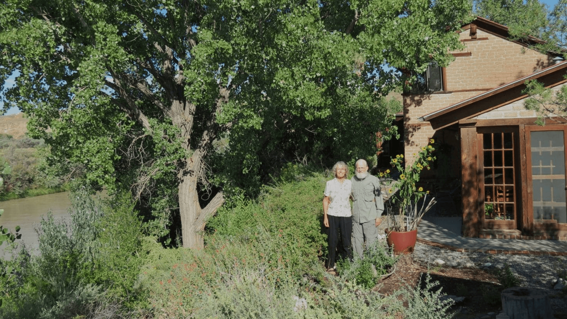 A couple stands together in front of a house surrounded by greenery near a river.