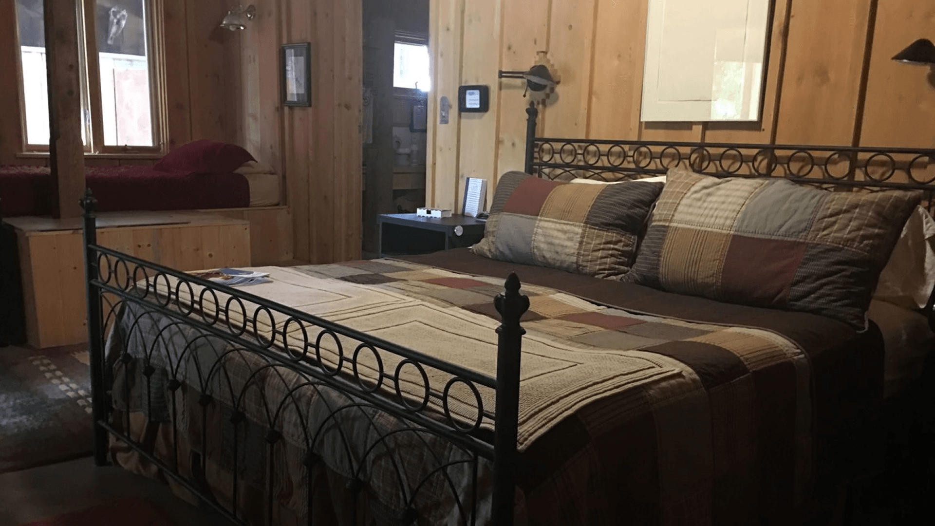A cozy bedroom featuring a metal bed frame with a patchwork quilt and wooden walls, with a second bed visible in the background.