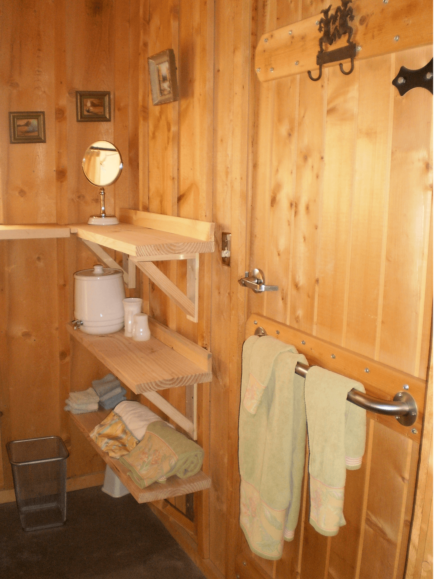 A wooden bathroom featuring shelves with towels, toiletries, and a mirror, alongside a door with a decorative handle.