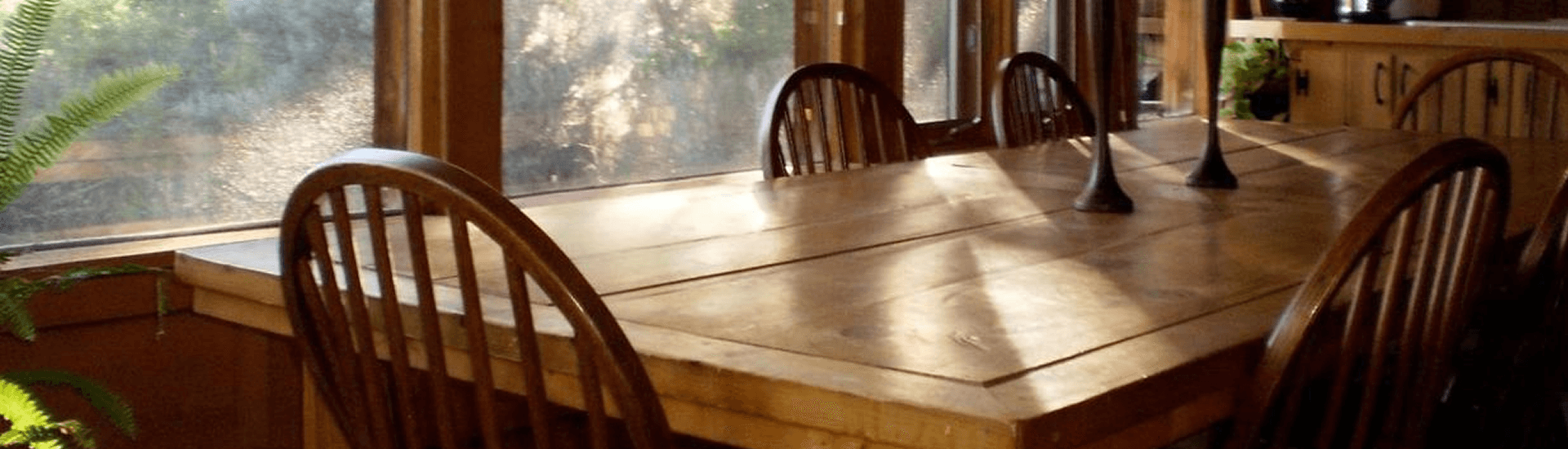 A wooden dining table surrounded by chairs, illuminated by natural light through large windows.